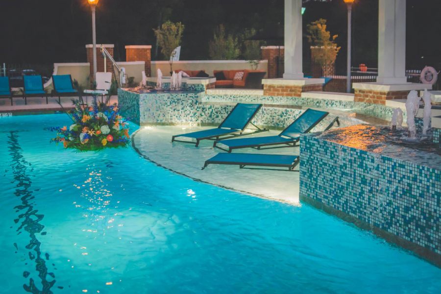 Attain at Quarterpath apartment homes with Two lounge chairs beside a glowing, tiled swimming pool at night with flowers and soft lighting.