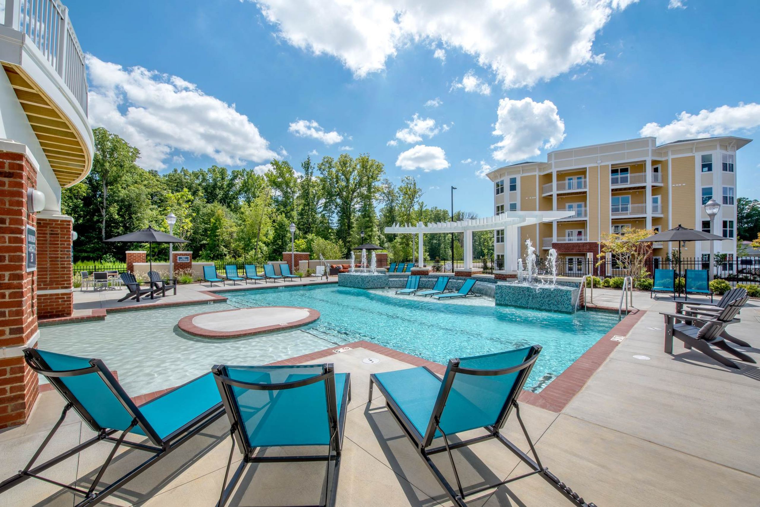 Attain at Quarterpath apartment homes with Outdoor pool with blue lounge chairs, water fountains, and apartment buildings in the background under a sunny sky.