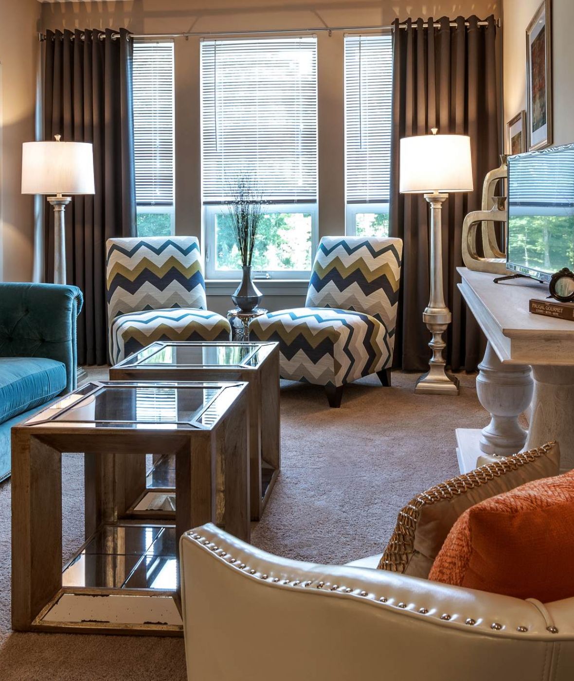 Attain at Quarterpath apartment homes with Modern living room with teal sofa, patterned chairs, glass coffee table, and bright natural light from windows.