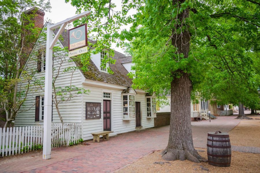 Attain at Quarterpath apartment homes with Historic white post office building with a sign, large tree, barrel, and picket fence along a brick path.