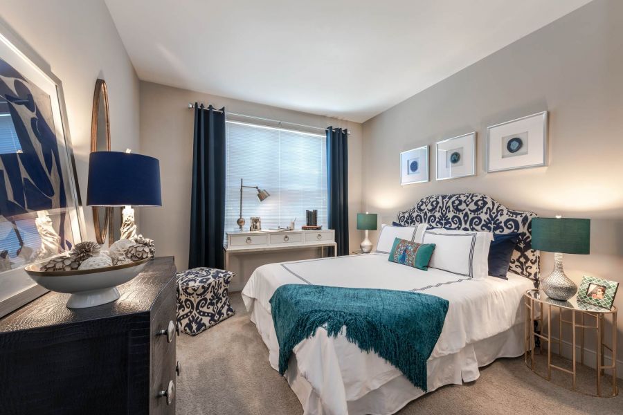 Attain at Quarterpath apartment homes with Elegant bedroom with patterned headboard, teal accents, lamps, desk, and framed art on beige walls.