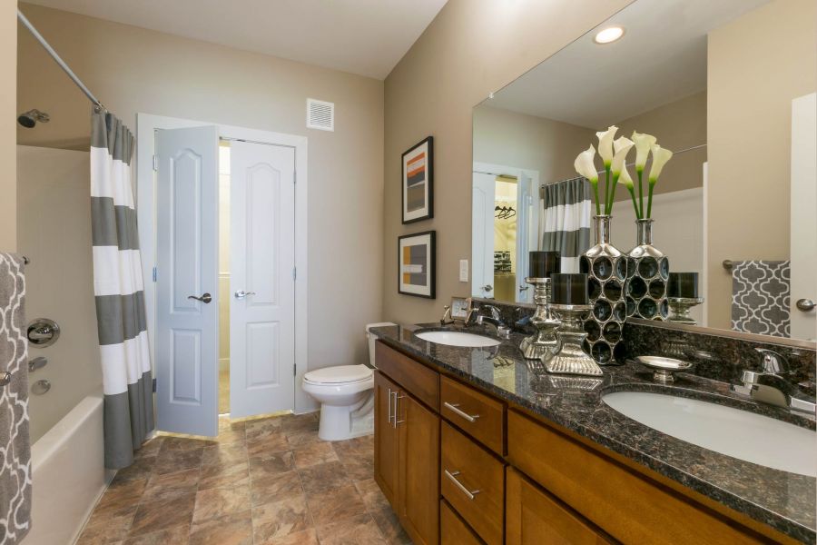 Attain at Quarterpath apartment homes with Modern bathroom with double sinks, granite countertop, and striped shower curtain, decorated with flowers.