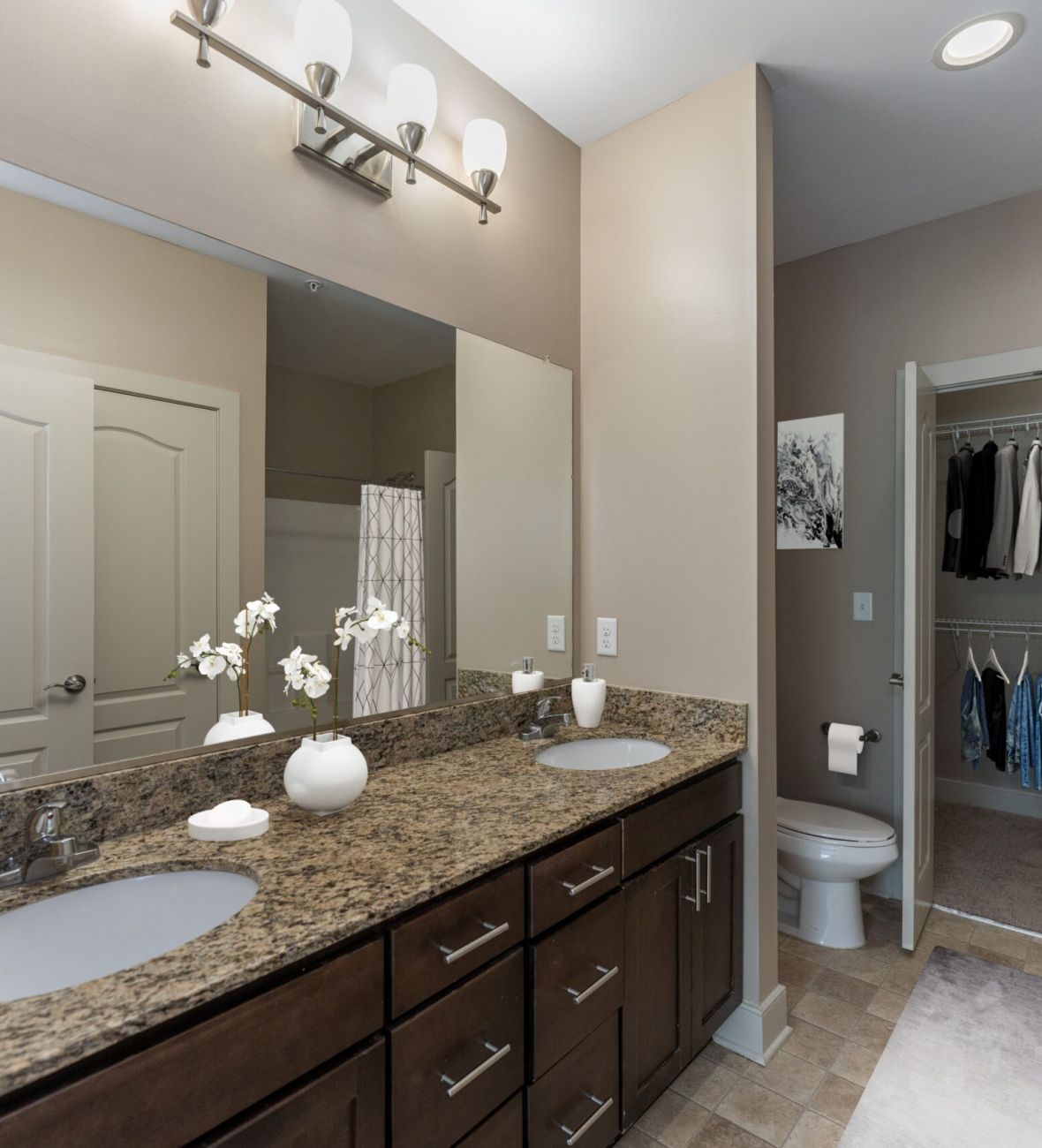 Attain on 5th apartment homes with A bathroom vanity with dark wood cabinets, a speckled granite countertop, double sinks, a large mirror, and an open walk-in closet in the background.