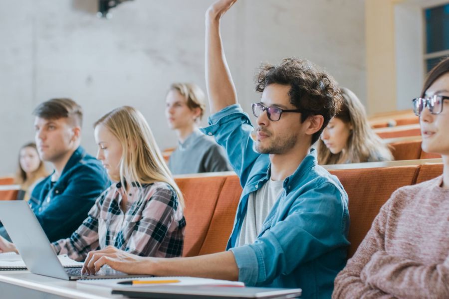 Attain on 5th apartment homes with Students sit in a lecture hall; one student raises his hand to ask a question.