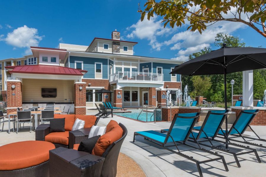 Attain at Quarterpath apartment homes with Outdoor pool area with lounge chairs, tables, and a modern building in the background under a blue sky.