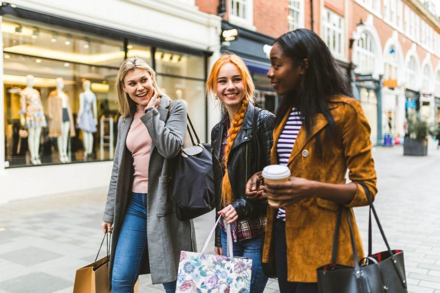 Attain at Quarterpath apartment homes with Three young women walk down a shopping street, smiling and carrying shopping bags and a coffee.