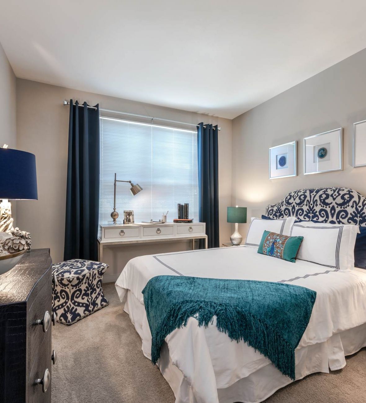 Attain at Quarterpath apartment homes with Elegant bedroom with patterned headboard, teal accents, lamps, desk, and framed art on beige walls.