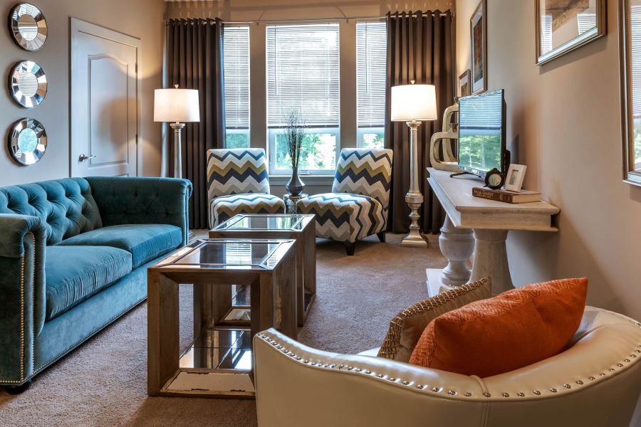 Attain at Quarterpath apartment homes with Modern living room with teal sofa, patterned chairs, glass coffee table, and bright natural light from windows.