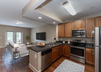 Attain at Quarterpath apartment homes with Modern kitchen with stainless steel appliances opens to a bright living room with a sofa, TV, and large windows.