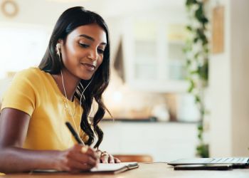 Attain on 5th apartment homes with Woman wearing earphones writes in a notebook while using a laptop at a table in a bright room with plants.